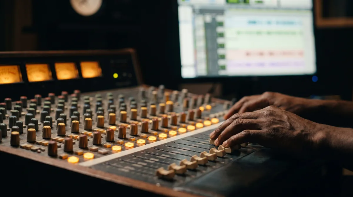 An engineer's hands on the faders of a large mixing console, the meters glowing amber
